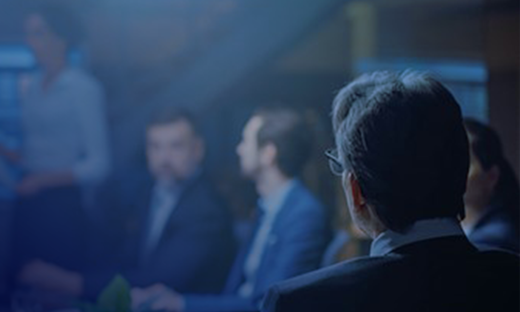 Une salle de réunion avec des personnes en tenue professionnelle, une personne mise en avant depuis l'arrière, les autres floues, en pleine conversation.