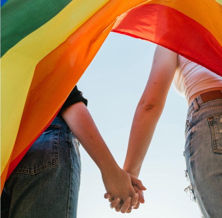 Dos personas cogidas de la mano bajo una bandera arcoíris del Orgullo, que simboliza la inclusión, la diversidad, la igualdad y el apoyo a los derechos humanos de las personas LGBTQ+.