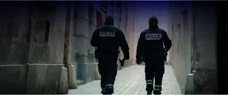 Deux policiers en uniforme sombre, avec l'inscription « POLICE » dans le dos, marchent dans une ruelle étroite et faiblement éclairée, bordée de grands immeubles. L'un des policiers tient un objet dans sa main droite. Le décor suggère un environnement urbain, mettant l'accent sur les thèmes de l'application de la loi et de la sécurité publique.