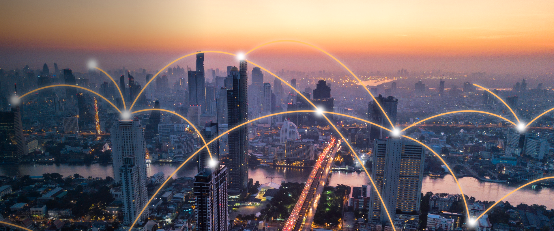 Horizon urbain au crépuscule avec des gratte-ciel reliés par des arcs lumineux, symbolisant la communication numérique et la connectivité. Une rivière et une autoroute traversent la ville, illuminées par les teintes chaudes orange et rose du ciel.