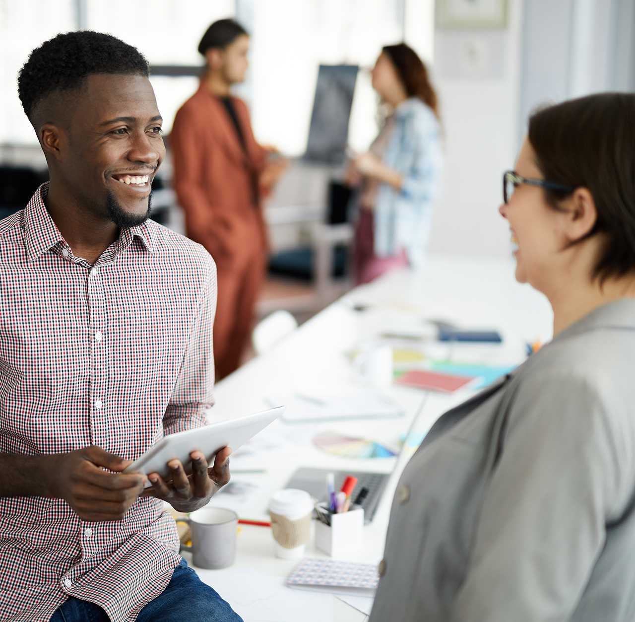 Dos estudiantes de pie junto a un escritorio en una oficina moderna, conversando. Una persona sostiene una tableta, mientras que sobre el escritorio se ven papeles, gráficos y material de oficina. Al fondo, otras personas interactúan cerca de una pizarra blanca, creando un ambiente de trabajo colaborativo.